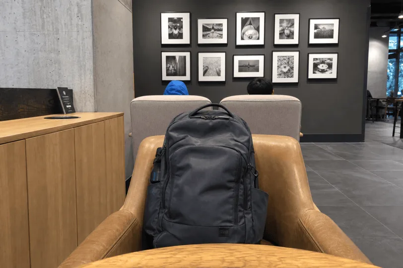 Webcam view from a MacBook on a café table — a backpack on the chair opposite, watched.